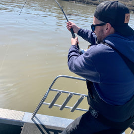 A man enthusiastically reeling in a catch during a White Sturgeon fishing charter on the Fraser River in Vancouver, British Columbia. He is wearing a blue hoodie, sunglasses, and a cap while standing on a boat.