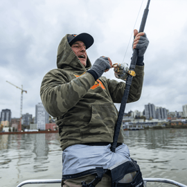 Excited man in camouflage hoodie holding a fishing rod at the back of a boat with the city in the background.